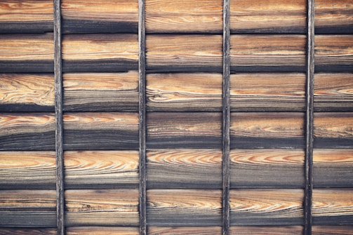 Wooden panels arranged in a horizontal pattern with visible grain texture and a weathered appearance. The wood showcases dark and light brown tones with blackened sections, suggesting intentional charring or aging.