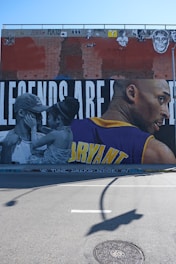 A large mural painted on a brick wall features a black and white image of a man and a child facing each other, with the child touching the man's face. On the right side, a colored portrait of an athlete is depicted wearing a yellow and purple jersey with the name 'BRYANT' visible. Above them, various graffiti and images are present on the brick wall.