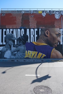 A large mural painted on a brick wall features a black and white image of a man and a child facing each other, with the child touching the man's face. On the right side, a colored portrait of an athlete is depicted wearing a yellow and purple jersey with the name 'BRYANT' visible. Above them, various graffiti and images are present on the brick wall.