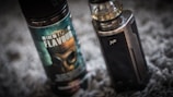 A vape device stands upright next to a bottle of vaping flavor liquid. The bottle features a label with bold text and an image of a gas mask. The vape device has a metallic finish with a clear tank on top.