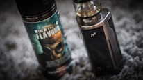 A vape device stands upright next to a bottle of vaping flavor liquid. The bottle features a label with bold text and an image of a gas mask. The vape device has a metallic finish with a clear tank on top.