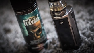 A vape device stands upright next to a bottle of vaping flavor liquid. The bottle features a label with bold text and an image of a gas mask. The vape device has a metallic finish with a clear tank on top.