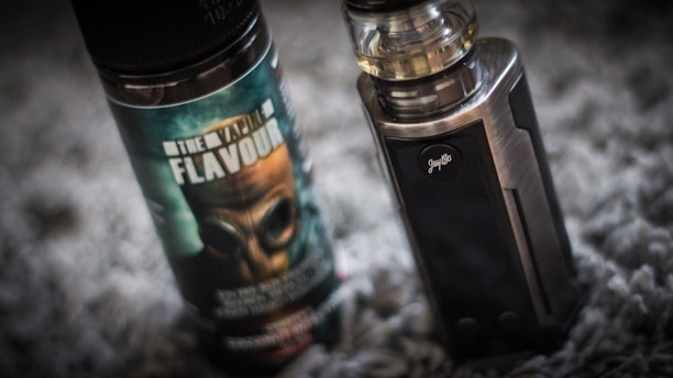 A vape device stands upright next to a bottle of vaping flavor liquid. The bottle features a label with bold text and an image of a gas mask. The vape device has a metallic finish with a clear tank on top.