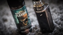A vape device stands upright next to a bottle of vaping flavor liquid. The bottle features a label with bold text and an image of a gas mask. The vape device has a metallic finish with a clear tank on top.