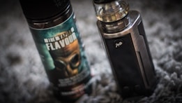 A vape device stands upright next to a bottle of vaping flavor liquid. The bottle features a label with bold text and an image of a gas mask. The vape device has a metallic finish with a clear tank on top.