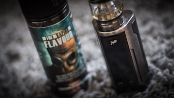 A vape device stands upright next to a bottle of vaping flavor liquid. The bottle features a label with bold text and an image of a gas mask. The vape device has a metallic finish with a clear tank on top.
