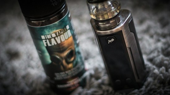 A vape device stands upright next to a bottle of vaping flavor liquid. The bottle features a label with bold text and an image of a gas mask. The vape device has a metallic finish with a clear tank on top.