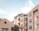 A group of modern apartment buildings with flat roofs and large windows. The buildings are constructed with beige-colored concrete or stucco, and feature glass-walled balconies or porches. A tree is visible in the foreground, and the sky is partly cloudy with a light blue shade.