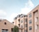 A group of modern apartment buildings with flat roofs and large windows. The buildings are constructed with beige-colored concrete or stucco, and feature glass-walled balconies or porches. A tree is visible in the foreground, and the sky is partly cloudy with a light blue shade.