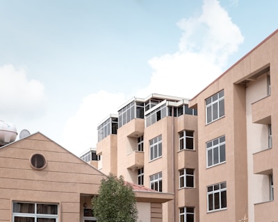 A group of modern apartment buildings with flat roofs and large windows. The buildings are constructed with beige-colored concrete or stucco, and feature glass-walled balconies or porches. A tree is visible in the foreground, and the sky is partly cloudy with a light blue shade.