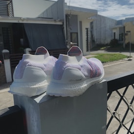 A pair of white and pastel-colored running shoes is placed on a concrete ledge. The shoes have a knitted design and the brand name 'ultra boost' is visible. The background shows a residential area with modern houses under a partly cloudy sky.