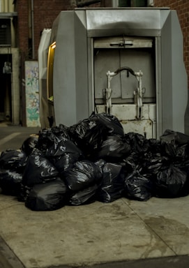 A large pile of black garbage bags is placed on a pavement next to a metal compactor. The setting appears to be an alleyway with brick walls and some street art visible in the background.