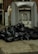 A large pile of black garbage bags is placed on a pavement next to a metal compactor. The setting appears to be an alleyway with brick walls and some street art visible in the background.