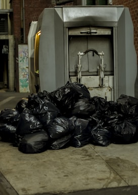 A large pile of black garbage bags is placed on a pavement next to a metal compactor. The setting appears to be an alleyway with brick walls and some street art visible in the background.