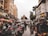 A bustling street scene features numerous motorbikes parked and people walking along a wet road flanked by tall buildings. Vendors with colorful umbrellas line the street on one side, and in the background, there is a church-like building with a distinctive architectural style.
