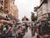 A bustling street scene features numerous motorbikes parked and people walking along a wet road flanked by tall buildings. Vendors with colorful umbrellas line the street on one side, and in the background, there is a church-like building with a distinctive architectural style.
