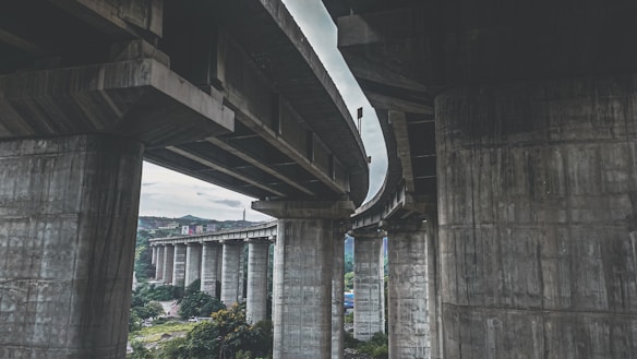 Massive concrete pillars support a complex overpass structure, with multiple layers of highway curving above. Surrounding the overpass are green hills and trees, indicating a natural environment that contrasts with the industrial nature of the bridge.