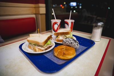 A table in a diner features a blue tray holding two sandwiches with lettuce, a hoagie roll, and sprouts. Also on the tray are a donut and a chocolate-covered pastry. Two drinks in red and white cups with straws are placed alongside a sugar dispenser.
