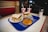 A table in a diner features a blue tray holding two sandwiches with lettuce, a hoagie roll, and sprouts. Also on the tray are a donut and a chocolate-covered pastry. Two drinks in red and white cups with straws are placed alongside a sugar dispenser.