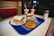 A table in a diner features a blue tray holding two sandwiches with lettuce, a hoagie roll, and sprouts. Also on the tray are a donut and a chocolate-covered pastry. Two drinks in red and white cups with straws are placed alongside a sugar dispenser.