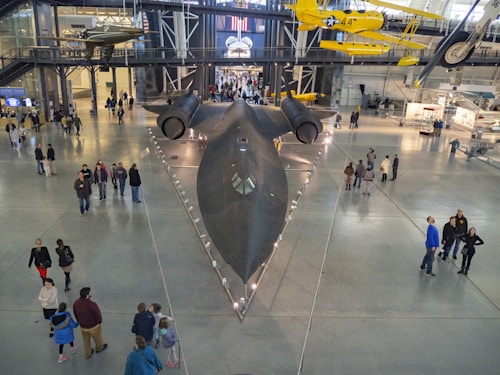 A large black aircraft is displayed indoors in a spacious museum setting, surrounded by a group of visitors. Several smaller, vintage aircraft are suspended from the ceiling above the main exhibit. The floor is gray, and the ceiling is high, with visible structural beams and lighting.
