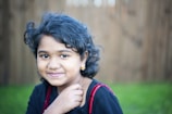 A young girl with curly hair is smiling gently, wearing a black outfit with red straps and small gold earrings. The background is softly blurred with hints of green and brown, suggesting an outdoor setting.