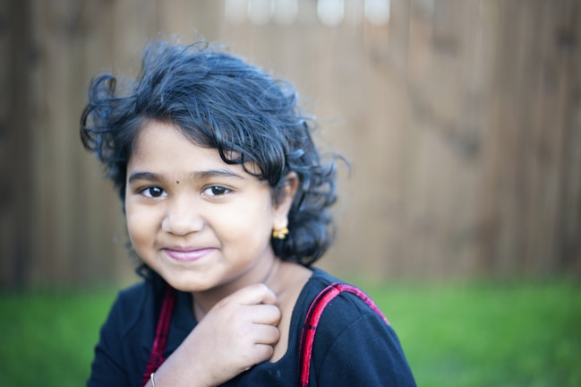 A young girl with curly hair is smiling gently, wearing a black outfit with red straps and small gold earrings. The background is softly blurred with hints of green and brown, suggesting an outdoor setting.