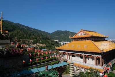 A large traditional Chinese building with an ornate yellow roof is situated in a picturesque mountainous area. The building is surrounded by vibrant red lanterns and colorful decorations. In the background, lush green hills rise under a clear blue sky.