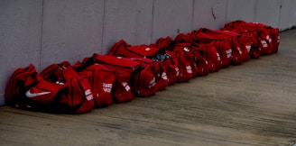 A row of red duffel bags placed side by side on a wooden floor against a concrete wall. Each bag seems identical in size and color, with some markings or text on them.
