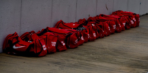 A row of red duffel bags placed side by side on a wooden floor against a concrete wall. Each bag seems identical in size and color, with some markings or text on them.