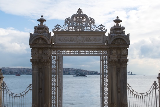 An ornate, historic gate with intricate carvings stands in the foreground, flanked by tall pillars. Beyond the gate, a large body of water stretches into the horizon, with a view of urban buildings and structures along the shoreline. The sky is partly cloudy, creating a serene and balanced atmosphere.
