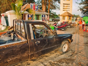 A weathered black pickup truck is partially sunk in sand near a roadside shop surrounded by tropical trees. Nearby, several motorbikes are parked, and people walk by, some dressed in light summer clothes. Signs and colorful decorations adorn the shop, and a small tower structure stands in the background, with the sun casting a warm glow over the scene.