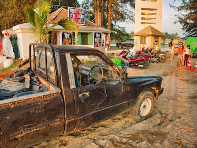 A weathered black pickup truck is partially sunk in sand near a roadside shop surrounded by tropical trees. Nearby, several motorbikes are parked, and people walk by, some dressed in light summer clothes. Signs and colorful decorations adorn the shop, and a small tower structure stands in the background, with the sun casting a warm glow over the scene.