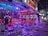 A vibrant, neon-lit bar scene featuring multiple high tables and stools, with a few patrons sitting and standing. The interior is adorned with bright lights and a decorative ceiling. Signs indicate the bar's name in glowing letters, and there's a lively, inviting atmosphere with colorful lighting.