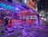 A vibrant, neon-lit bar scene featuring multiple high tables and stools, with a few patrons sitting and standing. The interior is adorned with bright lights and a decorative ceiling. Signs indicate the bar's name in glowing letters, and there's a lively, inviting atmosphere with colorful lighting.