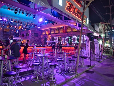 A vibrant, neon-lit bar scene featuring multiple high tables and stools, with a few patrons sitting and standing. The interior is adorned with bright lights and a decorative ceiling. Signs indicate the bar's name in glowing letters, and there's a lively, inviting atmosphere with colorful lighting.