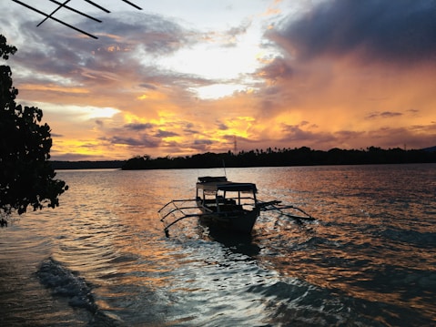 A traditional outrigger boat floats on a calm body of water during a vibrant sunset. The sky is filled with a mix of orange, yellow, and purple hues, reflecting on the water's surface. To the left, lush green foliage is visible, adding a natural frame to the scene.