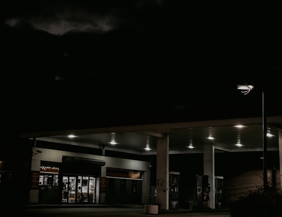 A dimly lit gas station at night with a convenience store visible in the background. The lighting highlights the empty fuel pumps and dark surroundings, creating a mysterious ambiance.