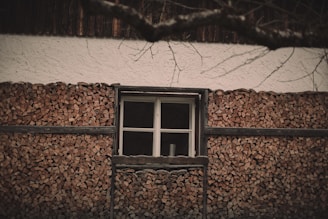 A wall of stacked firewood with a window in the center. Above the firewood, the building has a white plaster exterior. Tree branches, bare of leaves, are visible in the foreground, adding a natural element.