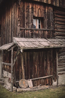 A rustic, weathered wooden building with a small window. The structure appears aged, with rough-hewn timber and an overhanging roof. There's a collection of wooden planks and a round wooden object on the ground. The grass in front contrasts with the dark wood.