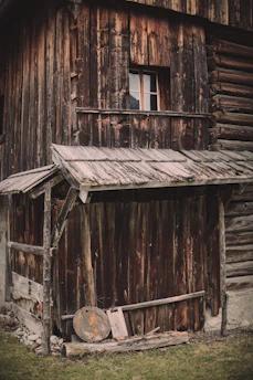 A rustic, weathered wooden building with a small window. The structure appears aged, with rough-hewn timber and an overhanging roof. There's a collection of wooden planks and a round wooden object on the ground. The grass in front contrasts with the dark wood.