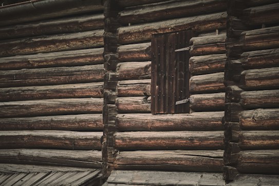 A rustic wooden cabin wall composed of horizontal logs stacked upon each other. The logs have a weathered appearance with a dark brown finish. A small wooden shuttered window is present, adding to the traditional aesthetic.
