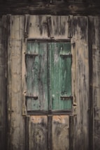 A weathered wooden wall with a rustic green-painted window shutter. The wood appears aged with visible textures and grain patterns. The shutter has a distressed look with faded paint and visible knots and imperfections.