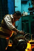 A man working intently on a large motor or engine in a dimly lit workshop. The environment is industrial, with tools and equipment visible on a workbench. Warm lighting highlights the man's focused expression and the metallic parts of the machinery.