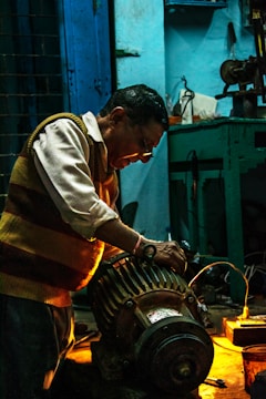 A man working intently on a large motor or engine in a dimly lit workshop. The environment is industrial, with tools and equipment visible on a workbench. Warm lighting highlights the man's focused expression and the metallic parts of the machinery.