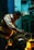 A man working intently on a large motor or engine in a dimly lit workshop. The environment is industrial, with tools and equipment visible on a workbench. Warm lighting highlights the man's focused expression and the metallic parts of the machinery.