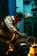 A man working intently on a large motor or engine in a dimly lit workshop. The environment is industrial, with tools and equipment visible on a workbench. Warm lighting highlights the man's focused expression and the metallic parts of the machinery.
