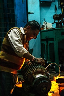 A man working intently on a large motor or engine in a dimly lit workshop. The environment is industrial, with tools and equipment visible on a workbench. Warm lighting highlights the man's focused expression and the metallic parts of the machinery.