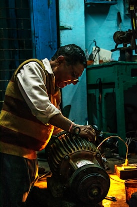 A man working intently on a large motor or engine in a dimly lit workshop. The environment is industrial, with tools and equipment visible on a workbench. Warm lighting highlights the man's focused expression and the metallic parts of the machinery.
