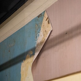 A section of wallpaper is peeling away from the wall, revealing an older, worn surface underneath with layers of blue and beige paint. The texture of the wallpaper is visible with some discoloration and possible mold spots.